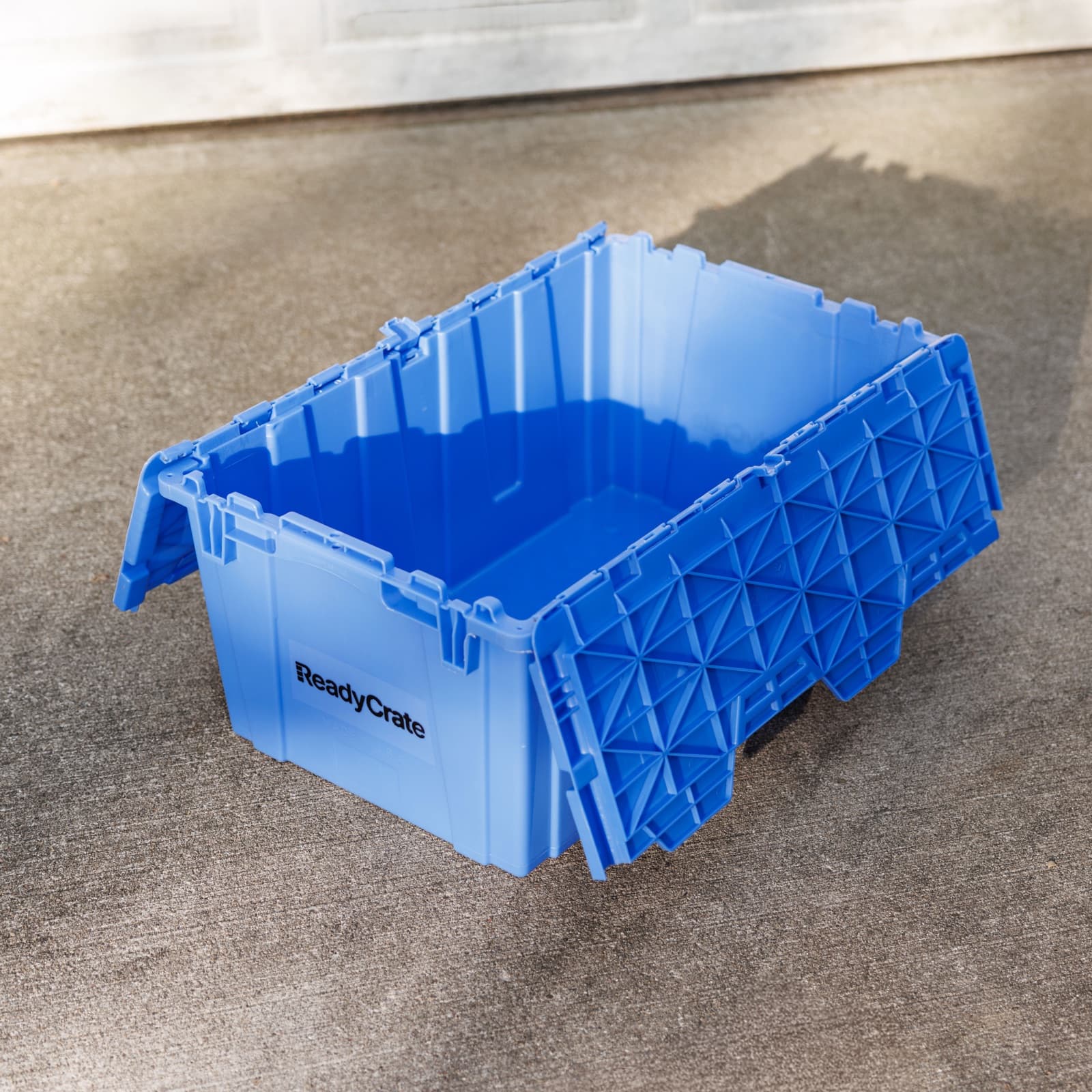 An open ReadyCrate tote on a driveway, lid folded back, with the attached lid and side handles visible.